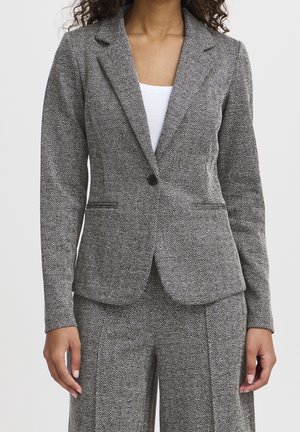 Vrouw die een aansluitend grijs visgraatcolbert en bijpassende broek draagt met een wit shirt eronder, staand tegen een effen achtergrond.
