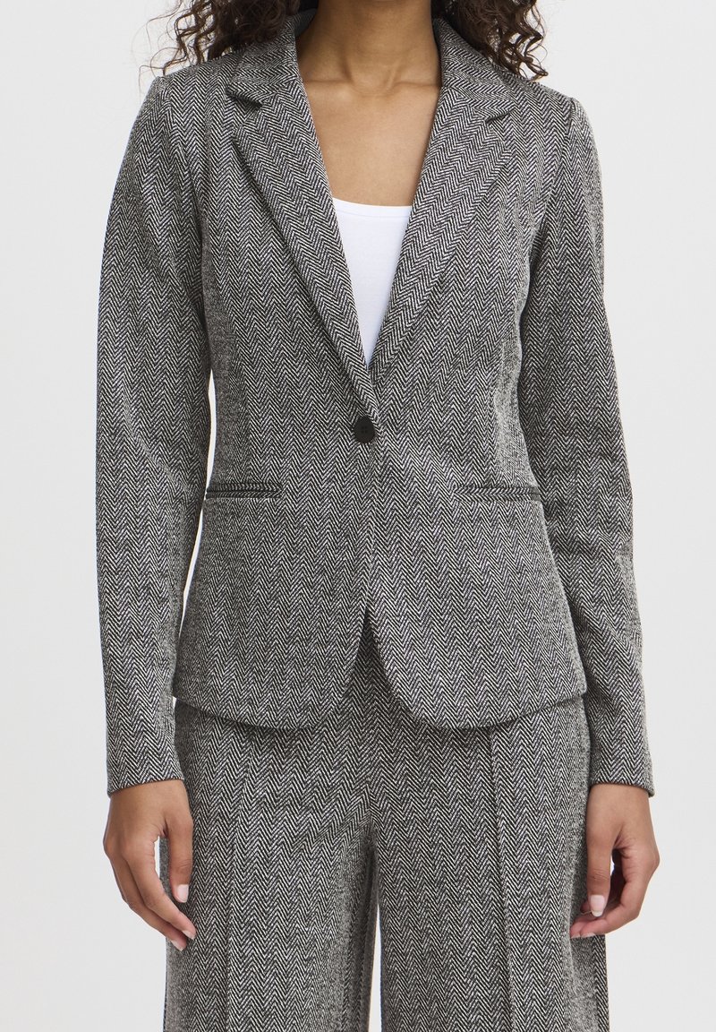 Femme portant un blazer gris en chevrons cintré et un pantalon assorti avec un top blanc en dessous, debout devant un fond uni.