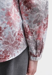 Chemise à manches longues florale en tissu léger avec des motifs rouges et gris, dotée de poignets boutonnés et d'une coupe décontractée. Détails texturés sur toute la surface.