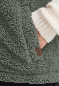 Veste en polaire verte avec un extérieur doux et texturé ; présente un intérieur en tricot beige et une petite étiquette en cuir pour le marquage.