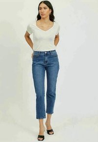 T-shirt branco de decote em V, jeans azul de cintura alta, comprimento cropped. Modelo usa sapatos de salto abertos pretos, de pé com as mãos na cintura, fundo neutro.