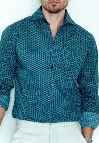 Man draagt een teal gekleurde button-down met patroon en opgerolde mouwen, en witte broek, staand tegen een witte muur.