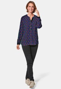 Langarmhemd van donkere stof met kleurrijk abstract patroon, met een V-hals en knoopdetails; gecombineerd met zwarte broek en sneakers.