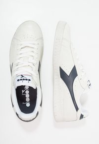 Vita lädersneakers med marinblå detaljer, texturerad yta, perforeringar på ovandelen, platta snören och texturerade gummisulor.