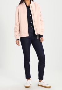 Giacca bomber rosa chiaro con chiusura a zip, polsini e orlo a coste, abbinata a una camicia blu navy a pois, jeans scuri e sneaker bianche.