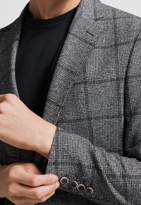 Szary marynarka w kratę z teksturowanej tkaniny, z klapami w kształcie szpicu i trzema guzikami na mankietach. Nosi się ją na czarnej koszuli.