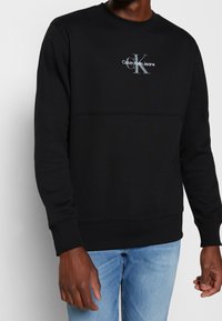 Svart sweatshirt i bomullsblandning med ribbad rund hals, långa ärmar och en diskret Calvin Klein-logotyp i grått på bröstet.