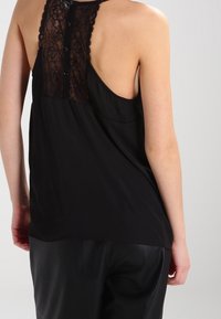 Svart ärmlös topp med spetsbakdel och knäppning, tillverkad av mjuk tyg som ger en avslappnad passform och mjuk känsla.