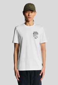 Weißes Baumwoll-T-Shirt mit kurzen Ärmeln, das auf der linken Brust ein schwarzes grafisches Design und einen runden Halsausschnitt aufweist. Das Modell trägt eine grüne Kappe.