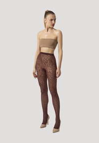 Bruna leggings med spetsmönster gjorda av genomskinligt material, i kombination med en nude bandeau-top och beigefärgade klackar, mot en neutral bakgrund.