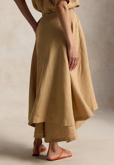 Moteris dėvi sluoksniuotą smėlio spalvos sijoną ir derančią palaidinę, stovinti basomis oranžinėse atviruose sandalėse ant neutralaus fono.