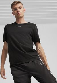 Čierne tričko s krátkym rukávom a okrúhlym výstrihom, na hrudi s bielym logom Puma, v kombinácii s čiernymi športovými nohavicami so zipsovými vreckami.