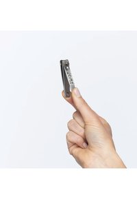 Main tenant un petit coupe-ongles en acier inoxydable avec des symboles gravés sur un fond blanc uni.