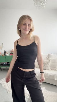 Νεαρή γυναίκα με ξανθά μαλλιά που φοράει μαύρο τοπ και παντελόνι στέκεται σε ένα φωτεινό σαλόνι με λευκό καναπέ και πράσινο ντουλάπι.