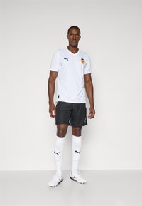 Hvit atletisk drakt med Valencia CF-logo, kombinert med svarte shorts, hvite sokker og hvite fotballsko; inkluderer logo aksenter på klærne.