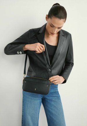 Kobieta w czarnej skórzanej kurtce i niebieskich dżinsach poprawiająca pasek czarnej torby na ramię na białym tle.