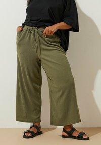Pantalons larges, légers et verts en tissu texturé, avec une taille à cordon de serrage, associés à des sandales plates noires avec un accent en boucle.