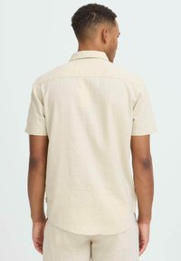 Chemise beige en lin à manches courtes avec un col étroit, un empiècement dans le dos et un ourlet arrondi. Design minimaliste, tissu texturé, coupe décontractée.