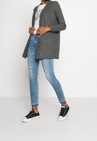 Cardigan gris abierto sobre una camiseta blanca con estampado, combinado con jeans ajustados de mezclilla clara rasgados y zapatillas de lona negras.