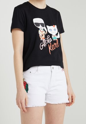 Black t-shirt with cartoon of man playing guitar and cat, text "Aloha Karl," paired with white denim shorts featuring floral embroidery.