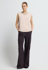 Frau, die ein beiges ärmelloses Oberteil mit einem mit Edelsteinen besetzten Ausschnitt und schwarze, weite Cargo-Hosen mit Taschen trägt, steht mit den Händen in den Taschen.