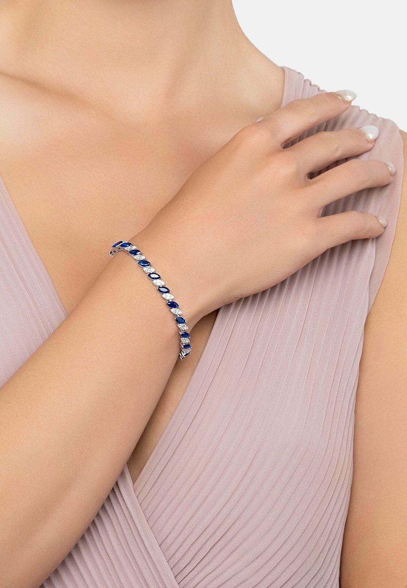 Silver bracelet featuring oval blue stones and clear accents, designed in a linked pattern, worn on a light-skinned arm against a soft fabric background.