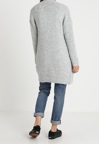 Cárdigan largo de punto gris claro con un ajuste holgado, dobladillo y puños acanalados, combinado con jeans azules enrollados y calzado negro.