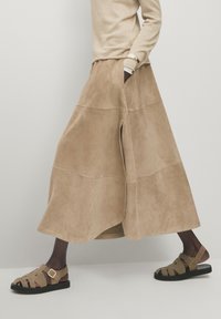 Jupe longue en daim beige avec des fentes latérales, complétée par des sandales marron à lanières avec une boucle et une semelle noire. Tissu à finition texturée.