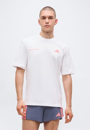 Młody mężczyzna w białym T-shircie z napisem "Running State of Mind" i granatowych sportowych szortach z różowymi paskami i logo, stojący na białym tle.