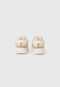 Sneaker beige e bianche con un corpo in mesh, colletto imbottito e suola in gomma testurizzata. Presentano strisce verticali e una linguetta sul retro.