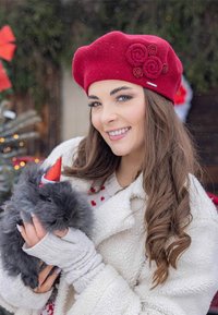 Berretto di lana rossa con fiori di stoffa e decorazioni, indossato con un cappotto di pile bianco. Tiene in braccio un coniglio nero e peloso con un piccolo cappello da festa rosso.