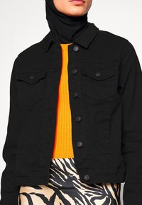 Personne portant un hijab noir, une veste en jean noire boutonnée par-dessus un pull orange vif, et une jupe beige avec de larges rayures noires zébrées.