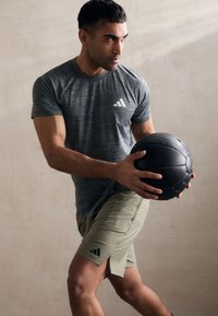 Grå atletisk t-shirt med stribet tekstur og hvidt logo, parret med olivengrønne shorts. Modellen holder en sort struktureret medicinbold.