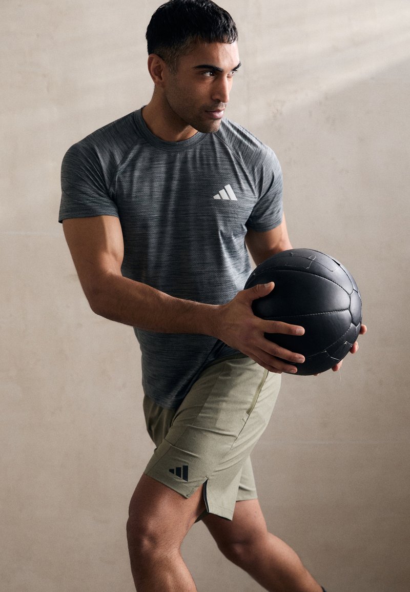Grå atletisk t-shirt med stribet tekstur og hvidt logo, parret med olivengrønne shorts. Modellen holder en sort struktureret medicinbold.