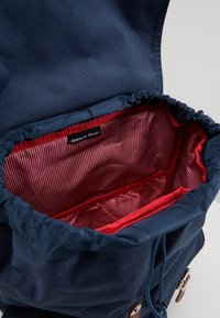 Granatowy plecak z tkaniny z wnętrzem w czerwono-białe paski. Posiada zapięcie na sznurek oraz wnętrzne kieszenie do organizacji.