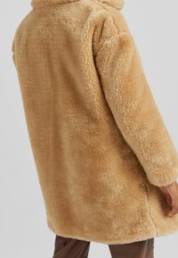 Manteau en fausse fourrure beige avec une surface douce et texturée, des manches longues et une coupe décontractée. Le manteau est doté d'un large col et d'une coupe mi-longue.