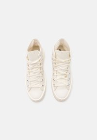 Paire de baskets montantes en toile couleur blanc cassé avec lacets, œillets métalliques et embouts en caoutchouc, disposées côte à côte sur un fond clair.