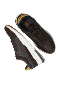 Bruine suede sneakers met een witte tussenzool en donkere accenten. Voorzien van geperforeerde patronen en een blauwe voering, aangevuld met gele merkvoering.