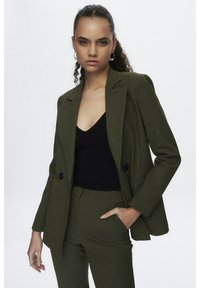 Groen geruit blazer met twee knopen, met notch-lapels en zakdetails, gedragen over een zwarte top. De broek heeft een bijpassend ruitpatroon.