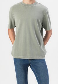 Joven adulto vistiendo una camiseta corta de manga suelta color verde oliva y jeans de mezclilla azules, de pie con las manos detrás de la espalda.