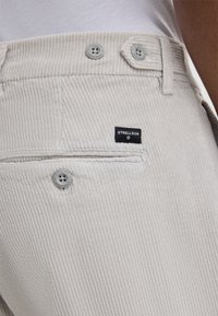 Pantaloni in corduroy grigio chiaro con una texture a righe, due bottoni in vita e un'etichetta nera con la scritta "STRELLSON" sulla tasca posteriore.