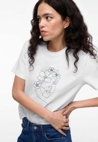 Witte t-shirt met een zwart lijntekening van een gezicht versierd met bloemelementen. Korte mouwen en een losse pasvorm, gecombineerd met denim jeans.