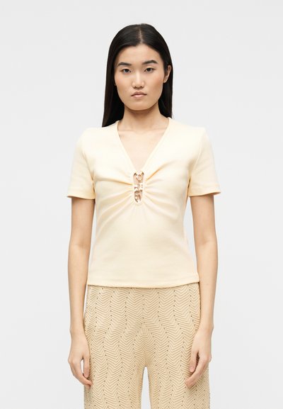 Femme aux cheveux raides et foncés portant un haut beige clair à manches courtes avec des détails à fermoirs métalliques et un pantalon beige texturé, debout devant un fond uni.