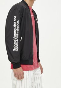 Giubbotto bomber nero con testo bianco "National Aeronautics and Space Administration" sulla manica, dotato di tasche con zip e polsini a coste.