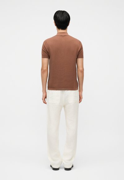 Homme portant un polo marron à manches courtes et un pantalon blanc ample, debout avec les bras détendus, de dos sur un fond blanc.