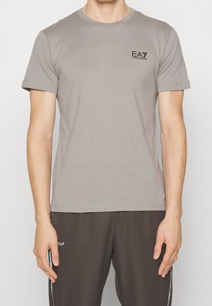 Homme portant un T-shirt manches courtes gris clair avec le logo "EA7 Emporio Armani" sur la poitrine et un pantalon foncé.