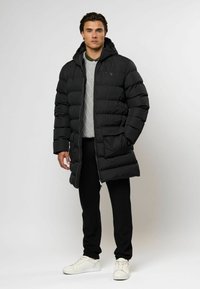 Mann iført en lang svart pufferjakke, grå genser, svarte bukser og hvite sneakers, står mot en lys ensfarget bakgrunn.