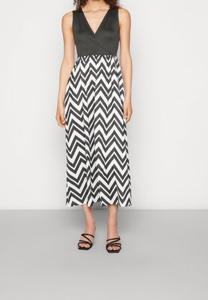 Femme portant une robe noire sans manches avec une jupe à motif zigzag noir et blanc et des sandales noires à brides, debout sur un sol gris.