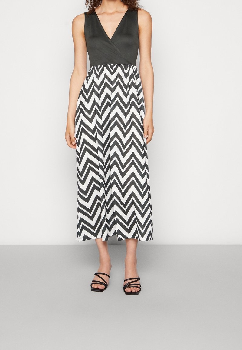 Femme portant une robe noire sans manches avec une jupe à motif zigzag noir et blanc et des sandales noires à brides, debout sur un sol gris.