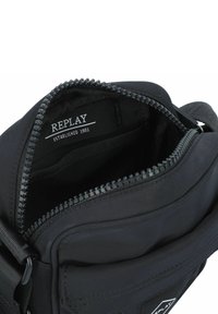 Schwarze Stofftasche mit Reißverschluss, innen mit einem Logotag, und einer glatten, matten Oberfläche. Verstellbarer Schulterriemen inklusive.